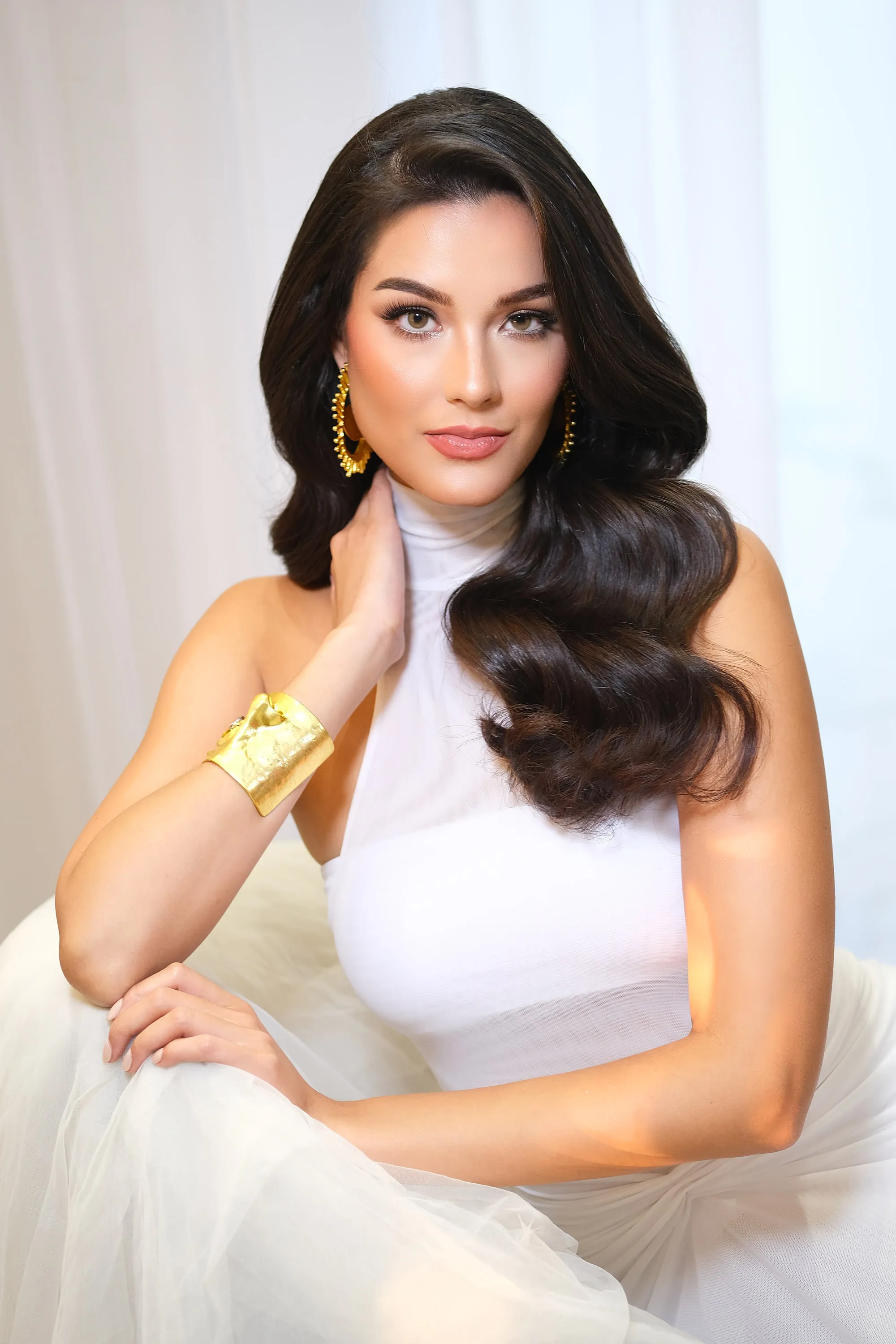 Editorial make-up with a warm sculpted eye and a nude lip, seated, with deep waves brought over one shoulder and a gold cuff at the wrist.