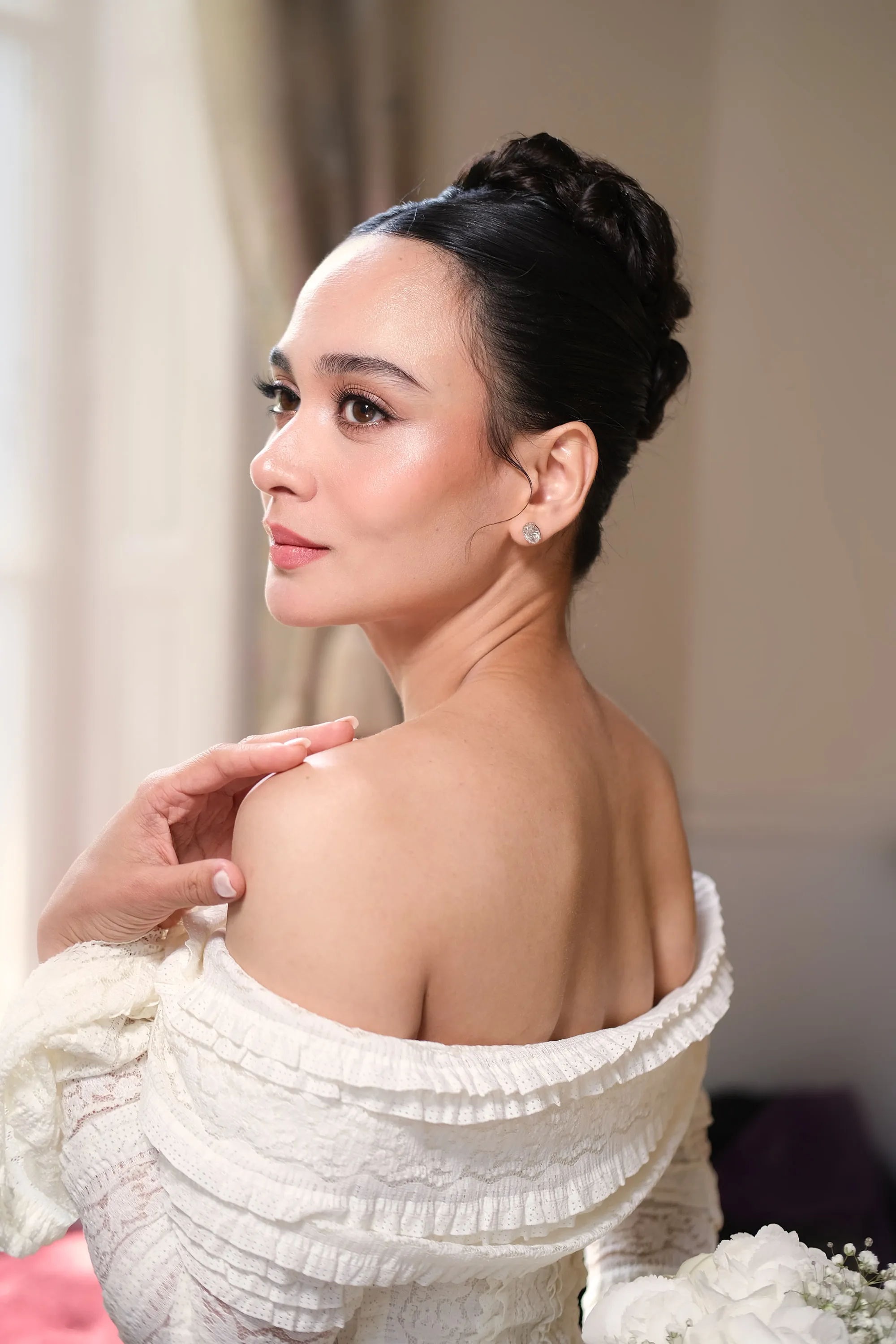 Editorial make-up seen in profile, with a lifted lash line and a warm cheek, worn with a high bun and a ruffled off-shoulder sleeve.