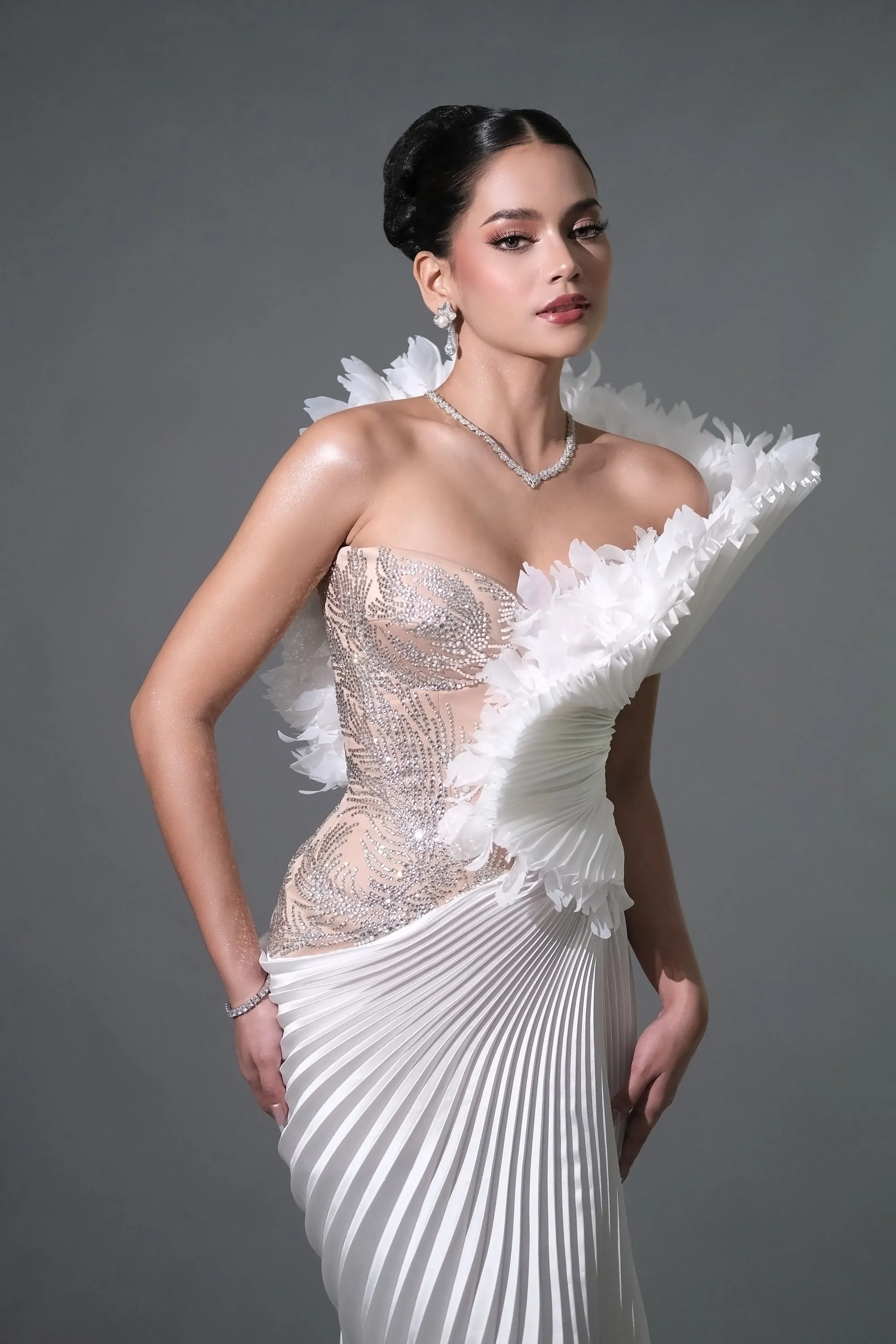 Full-length pageant look with a sculpted bronze eye and a high sleek bun, worn with a feathered and pleated ivory gown against a grey studio wall.